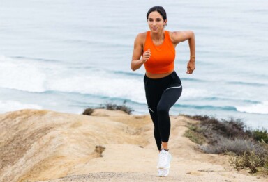Woman in athletic apparel running up side of mountain against ocean backdrop