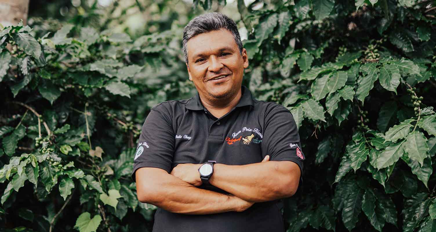 Photo of a coffee farmer standing with his arms crossed and a smile on his face in front of green foliage.