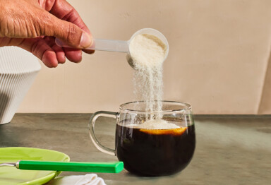 A hand pouring collagen powder into a cup of coffee on a counter.