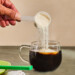 A hand pouring collagen powder into a cup of coffee on a counter.